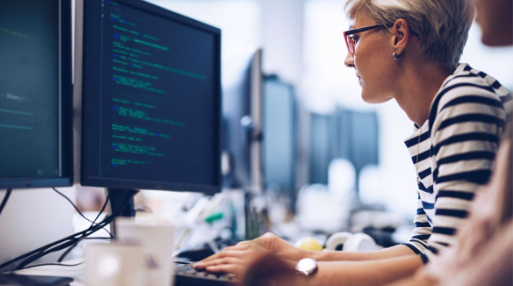 short-haired blonde woman with re glasses looks at code on desktop monitor